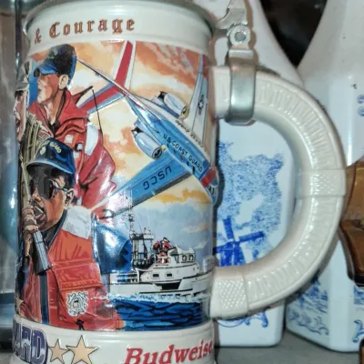 Sale Photo Thumbnail #60: Vintage Budweiser lidded beer stein honoring the United States Coast Guard, featuring colorful graphics of personnel, aircraft, and vessels.