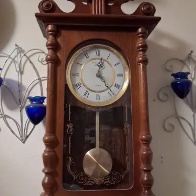 Sale Photo Thumbnail #66: Vintage Howard Miller quartz wall clock in a wooden case with a brass pendulum and Roman numeral dial, flanked by decorative metal wall sconces with blue glass inserts.