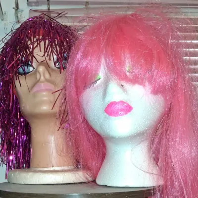 Sale Photo Thumbnail #22: Two mannequin heads displaying colorful wigs, featuring a metallic purple tinsel wig on a plastic head and a bright pink synthetic wig on a styrofoam head.