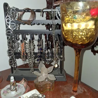 Sale Photo Thumbnail #32: Assorted costume jewelry collection, featuring necklaces, bracelets, and rings displayed on a black metal stand, alongside a decorative amber glass goblet and small trinket dishes.