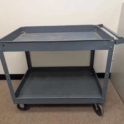 Sale Photo Thumbnail #239: Industrial gray metal utility cart with two deep shelves, a push handle, and caster wheels, showing some wear from use.