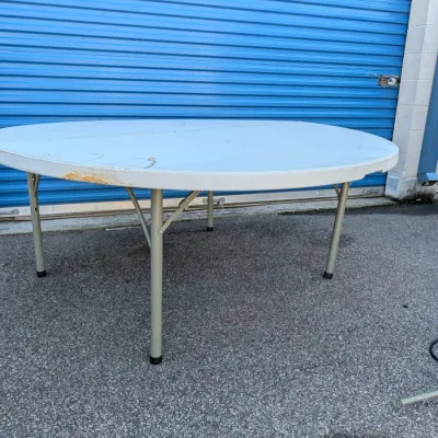 Sale Photo Thumbnail #23: Large round folding table with a white plastic top and metal legs, showing some wear and discoloration on the edge.