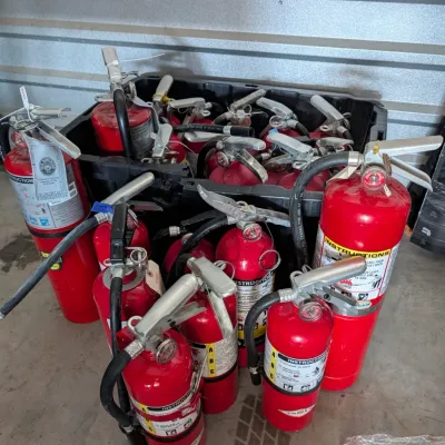 Sale Photo Thumbnail #22: A collection of used red fire extinguishers of various sizes, some stored in a black plastic bin.