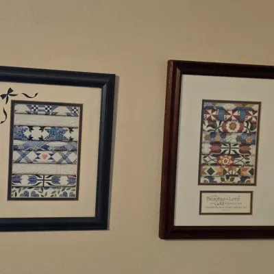 Pair of framed art prints depicting stacks of colorful quilts, one in a black frame with a ribbon detail and the other in a dark wood...