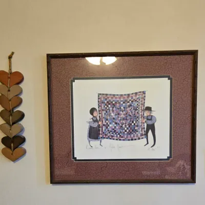 Framed P. Buckley Moss print depicting an Amish couple holding a quilt, flanked by rustic wooden heart wall hangings and inspirational...