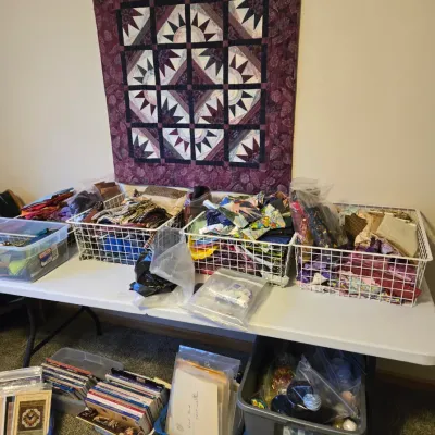 Handmade geometric quilt hanging on a wooden wall rack, displayed above a table filled with wire baskets of fabric scraps and quilting...