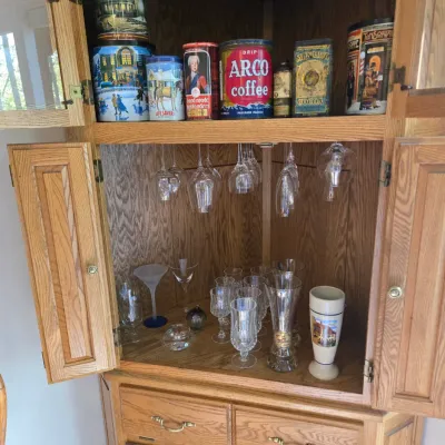 Sale Photo Thumbnail #67: Oak corner hutch featuring a collection of vintage advertising tins on the top shelf and various glassware, including wine glasses and tumblers, on the lower shelves.