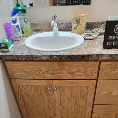 Sale Photo Thumbnail #66: Oak bathroom vanity with a laminate countertop, white drop-in sink, and chrome faucet, shown with various toiletries and a boxed Mirrorvana mirror.