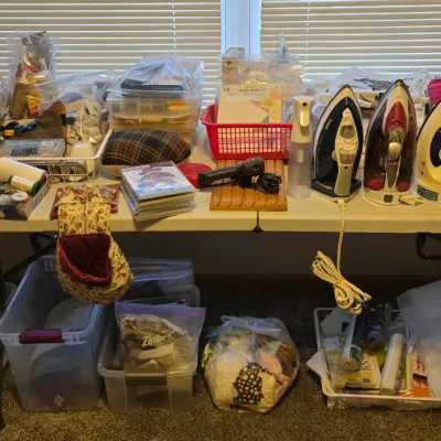 Assortment of sewing and crafting supplies, including three electric irons, thread spools, fabric scraps, and storage bins arranged on...