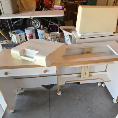 Sale Photo Thumbnail #104: White laminate sewing cabinet on casters with a drop-down platform, featuring a Husqvarna Viking sewing machine case resting on top.
