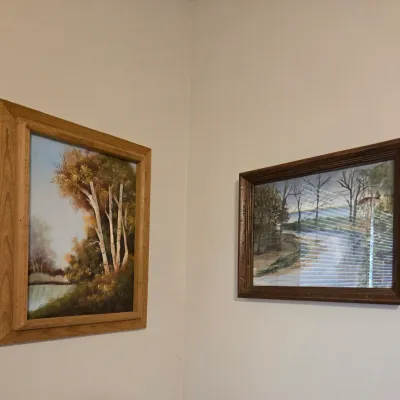 Sale Photo Thumbnail #39: Two framed landscape paintings, including an oil painting of birch trees by a river in a light oak frame and a glazed watercolor of a wooded path in a dark wood frame.
