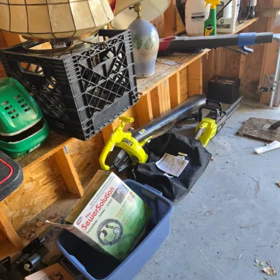 Sale Photo Thumbnail #100: Assorted garage items including a Ryobi leaf blower and chainsaw, a SewerSolution kit in its box, and various table lamps stored on a wooden shelf.