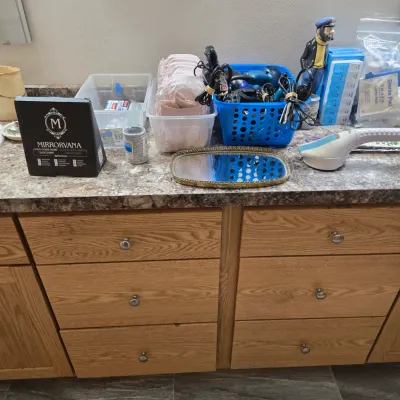 Sale Photo Thumbnail #65: Assorted personal care and medical supplies, including a Mirrorvana mirror in its box, adult diapers, hair dryers in a blue basket, and a handheld massager, arranged on a laminate countertop.