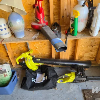 Sale Photo Thumbnail #96: Assorted lawn care equipment, including a Ryobi 40V cordless chainsaw and leaf blower with collection bag, alongside a red corded Toro blower vac and various household items on wooden shelving.
