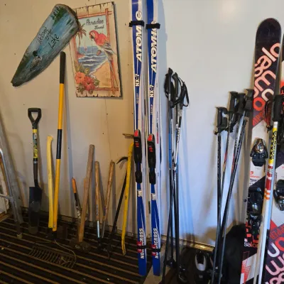 Sale Photo Thumbnail #87: Assorted outdoor gear including Atomic NX-7 cross-country skis, Salomon downhill skis, ski poles, and various hand tools leaning against a wall.