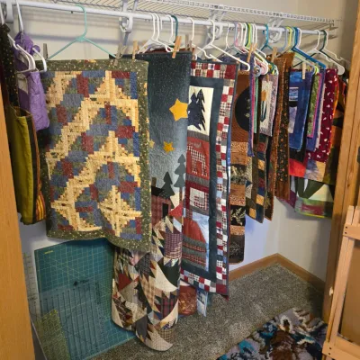 Collection of handmade quilts and wall hangings in various patterns and sizes, displayed on hangers in a closet.