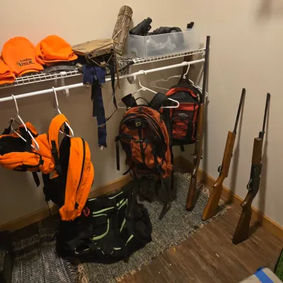 Assortment of hunting and outdoor gear, including three long guns, orange safety vests and hats, backpacks, and a duffel bag.
