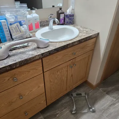 Sale Photo Thumbnail #64: Bathroom vanity with oak cabinetry, laminate countertop, and a white drop-in sink, shown with various medical supplies and a digital scale.