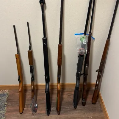 Collection of six long guns, including rifles and shotguns with wood and synthetic stocks, leaning against a wall.