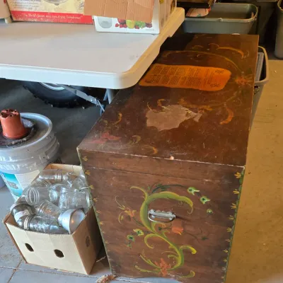 Sale Photo Thumbnail #82: Vintage hand-painted wooden trunk featuring floral rosemaling details and metal side handles, showing some surface wear and a paper label on top.
