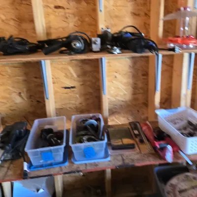 Sale Photo Thumbnail #109: Assorted power tools, including a corded drill and angle grinder, organized on wooden shelves alongside plastic storage bins containing hardware and accessories.