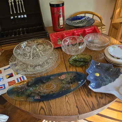 Sale Photo Thumbnail #68: Assortment of glassware and serving dishes, including cut glass bowls, a long art glass platter, a ceramic tree-shaped dish, and a boxed silverware set, displayed on a wooden dining table.