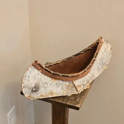 Sale Photo Thumbnail #58: Handcrafted miniature birch bark canoe model with stitched detailing, displayed on a rustic wooden pedestal stand.