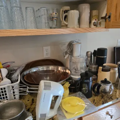 Sale Photo Thumbnail #70: Assorted kitchenware including a Hamilton Beach hand mixer, food processor, French press, mixing bowls, and glassware displayed on a countertop and cabinet shelves.
