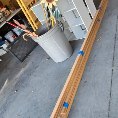 Sale Photo Thumbnail #94: Assorted garage items including bundles of wood trim secured with blue tape, a grey plastic trash can holding garden tools and a decorative metal sunflower, and a white laminate cube organizer.