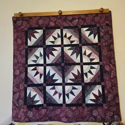 Handcrafted patchwork quilt wall hanging featuring a geometric starburst pattern in shades of purple, plum, and cream, displayed on a...