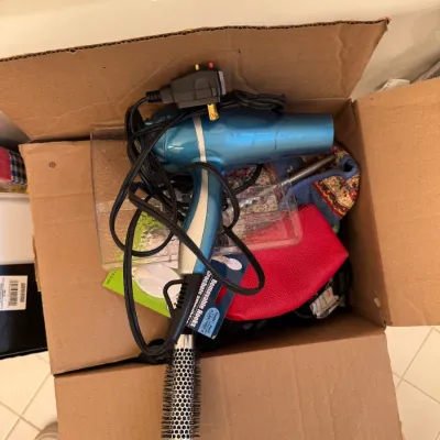 Sale Photo Thumbnail #207: Cardboard box containing a blue hair dryer, a round styling brush, a red pouch, and other miscellaneous household items.