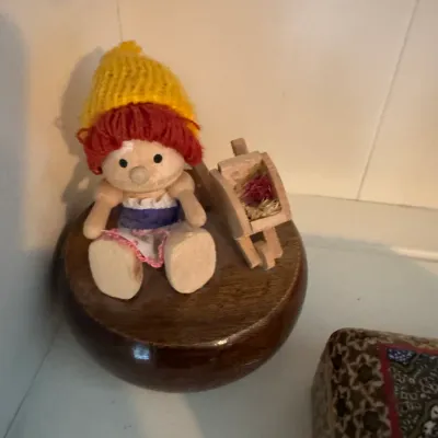 Sale Photo Thumbnail #182: Vintage wooden music box featuring a small doll with a yellow knit hat and red hair sitting next to a miniature wheelbarrow.