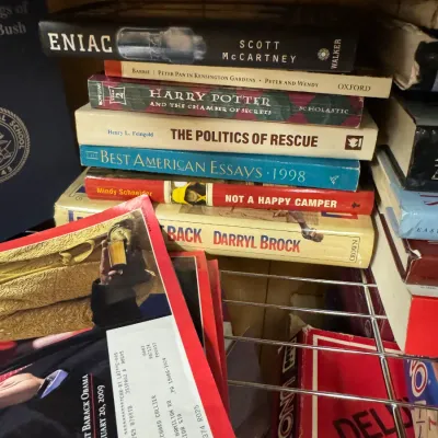 Sale Photo Thumbnail #120: A stack of assorted hardcover and paperback books, including "ENIAC" by Scott McCartney and "Harry Potter and the Chamber of Secrets," resting on a wire shelf alongside magazines and board games.