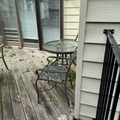 Sale Photo Thumbnail #338: Three-piece outdoor bistro set featuring a round glass-top table and two dark metal armchairs with woven strap seats, situated on a wooden deck.