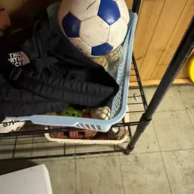 Sale Photo Thumbnail #149: Assorted sports equipment including a blue and white soccer ball, a black The North Face bag, and various smaller balls stored in plastic baskets on a wire shelf.