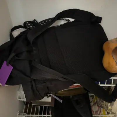 Sale Photo Thumbnail #163: Black backpack with padded shoulder straps and a purple luggage tag, resting on a wire shelf.