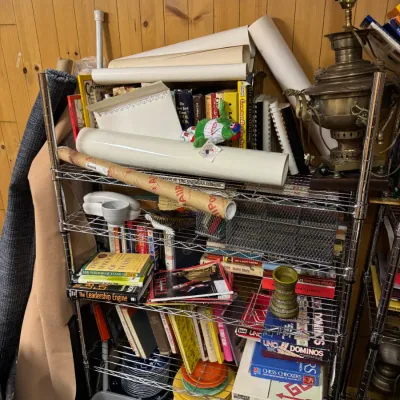 Sale Photo Thumbnail #130: Five-tier wire shelving unit filled with books, board games, rolled paper, and vintage collectibles, including a brass samovar lamp.
