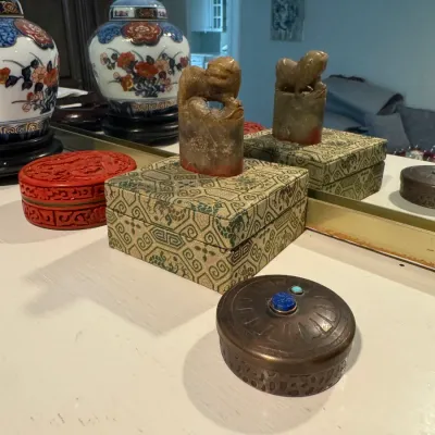 Sale Photo Thumbnail #256: Collection of Asian decorative items, including a carved stone seal with a foo dog finial on a patterned fabric box, a red cinnabar-style box, and a small metal trinket box with inset stones.