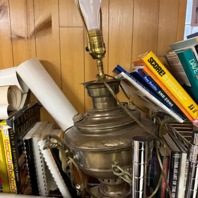 Sale Photo Thumbnail #123: Vintage brass samovar converted into a table lamp, showing some tarnish and wear, surrounded by books and paper rolls.