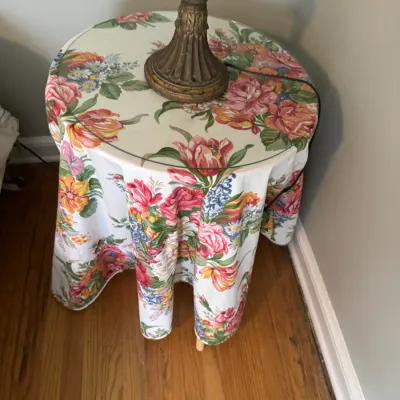 Sale Photo Thumbnail #204: Round accent table draped in a vibrant floral tablecloth, topped with a protective glass surface and a vintage-style table lamp.