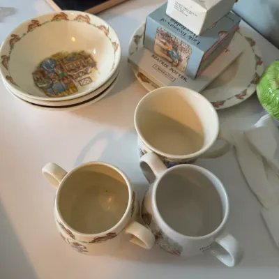 Sale Photo Thumbnail #215: Collection of vintage Beatrix Potter Peter Rabbit children's dishware, including bowls, mugs, and double-handled cups, alongside boxed gift sets.