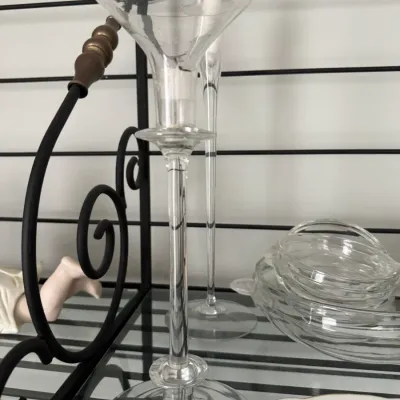 Sale Photo Thumbnail #348: Pair of tall, elegant clear glass candlesticks with flared rims, displayed alongside a stack of small glass dishes on a black metal baker's rack.