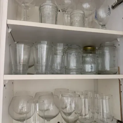 Sale Photo Thumbnail #333: Assorted glassware collection, including wine glasses, tumblers, and glass jars, arranged on white cabinet shelves.