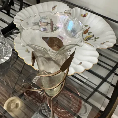 Sale Photo Thumbnail #340: Iridescent glass trumpet vase with a ruffled rim, supported by a brass-finished metal stand with leaf accents, displayed in front of a scalloped porcelain serving dish.