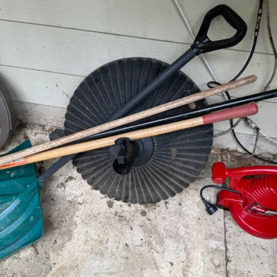 Sale Photo Thumbnail #74: Assorted outdoor items including a heavy black patio umbrella base, a green snow shovel, long-handled garden tools, and a red Toro electric leaf blower.