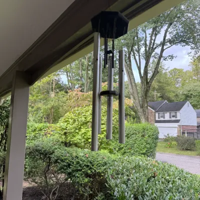 Sale Photo Thumbnail #308: Large metal wind chime with silver-toned tubes and a dark wooden top and clapper, hanging from a porch eave.