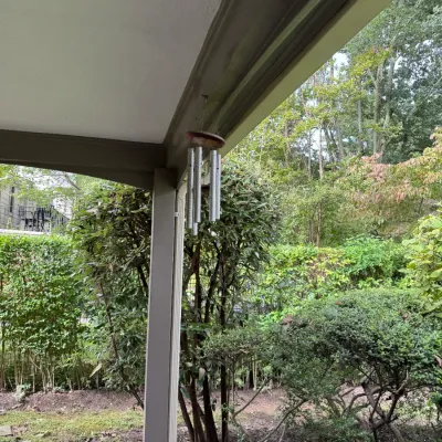 Sale Photo Thumbnail #309: Metal wind chime with a wooden top hanging from a porch eave, overlooking a lush green garden.