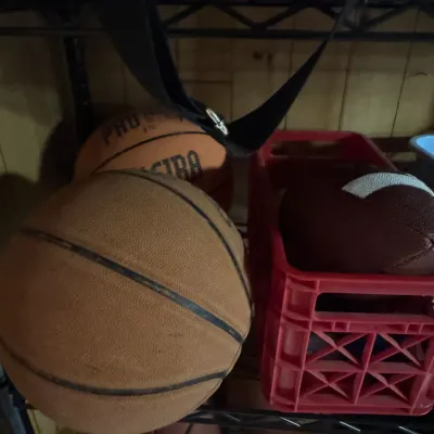 Sale Photo Thumbnail #147: Assorted sports balls, including worn leather basketballs and a football in a red plastic crate, stored on wire shelving.