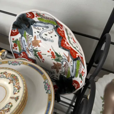 Sale Photo Thumbnail #344: Vintage Japanese Imari-style porcelain bowl with scalloped edges, featuring hand-painted floral and fish motifs in red, green, and blue, displayed alongside a stack of fine china plates.