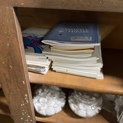 Sale Photo Thumbnail #105: A stack of vintage Maxwell House Haggadahs and other booklets on a wooden shelf, above glass jars filled with packing peanuts.
